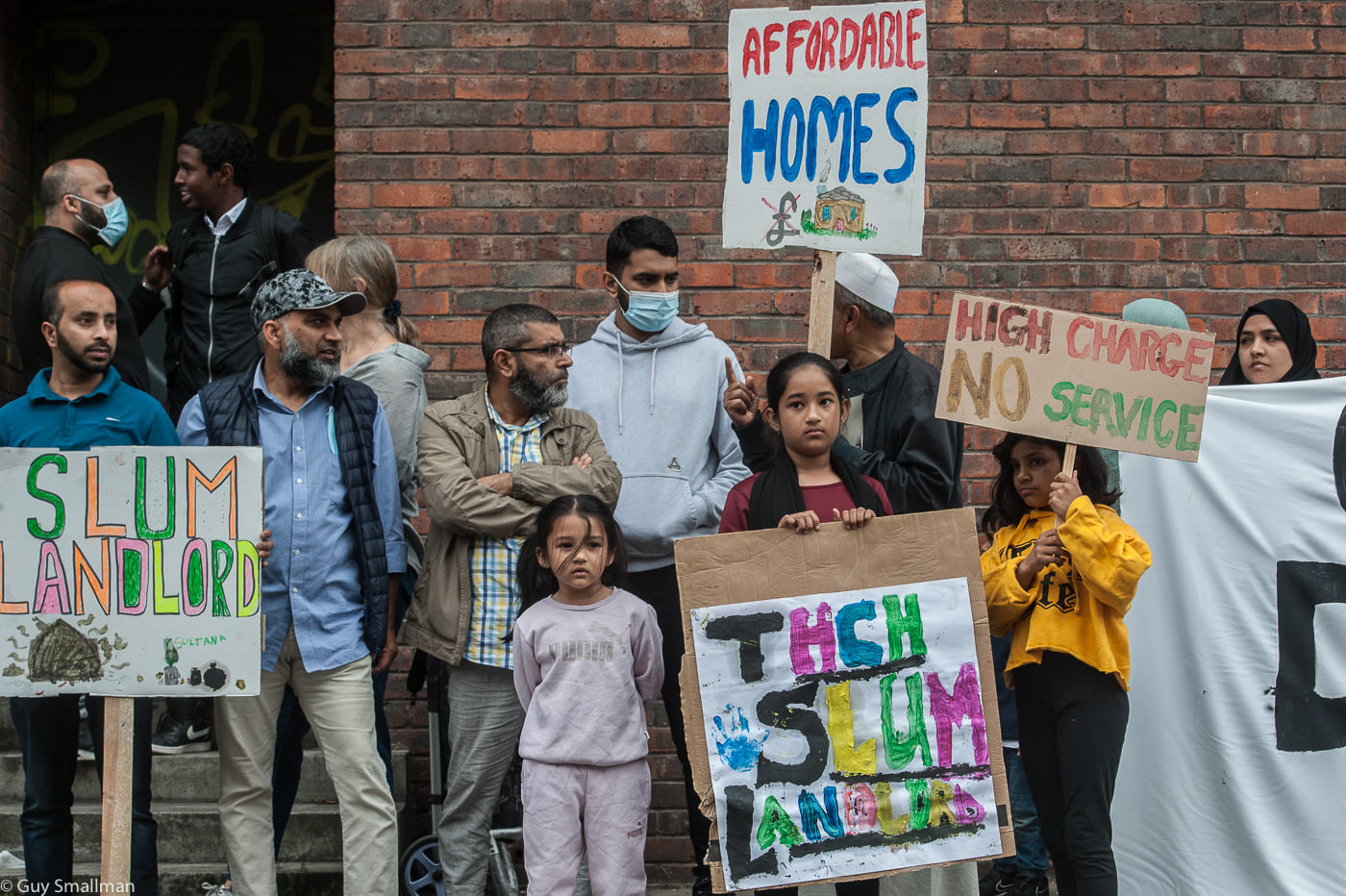 People stand in front of a building with placards that read 'Affordable Homes' and 'scum landlord'