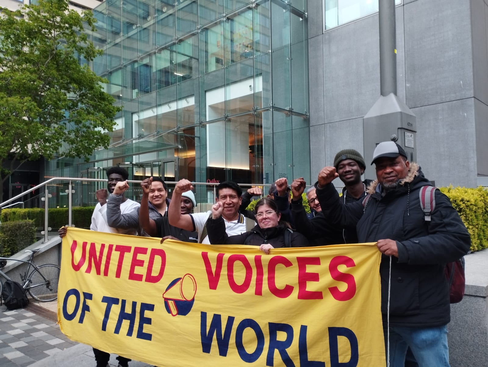 People stand with with fists in the air behind a banner that reads UNITED VOICES OF THE WORLD